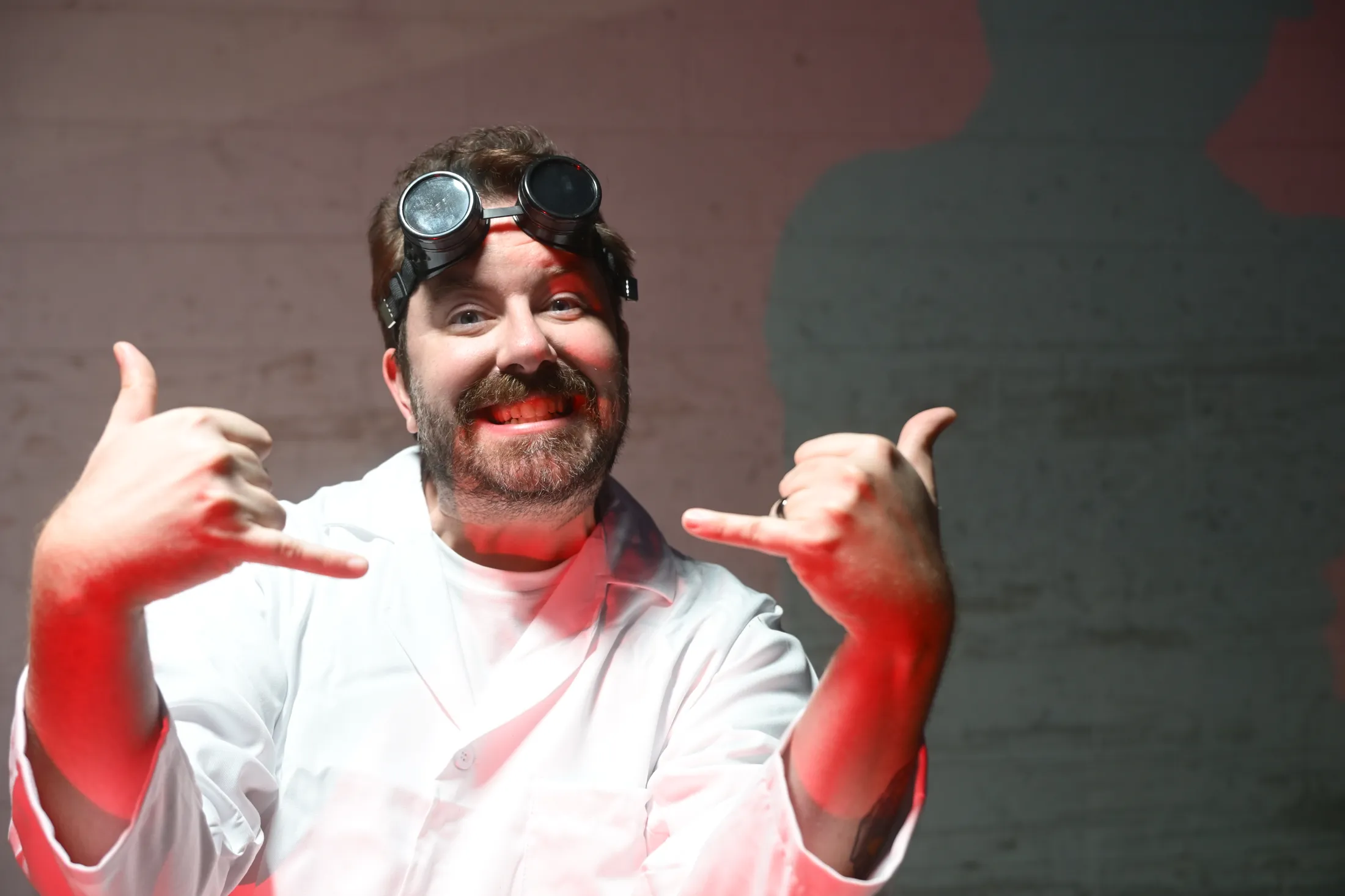 Mad scientist in a lab coat and goggles grinning and throwing shakas in red light, from Rusty Rehl's Combat Sport Supply ad