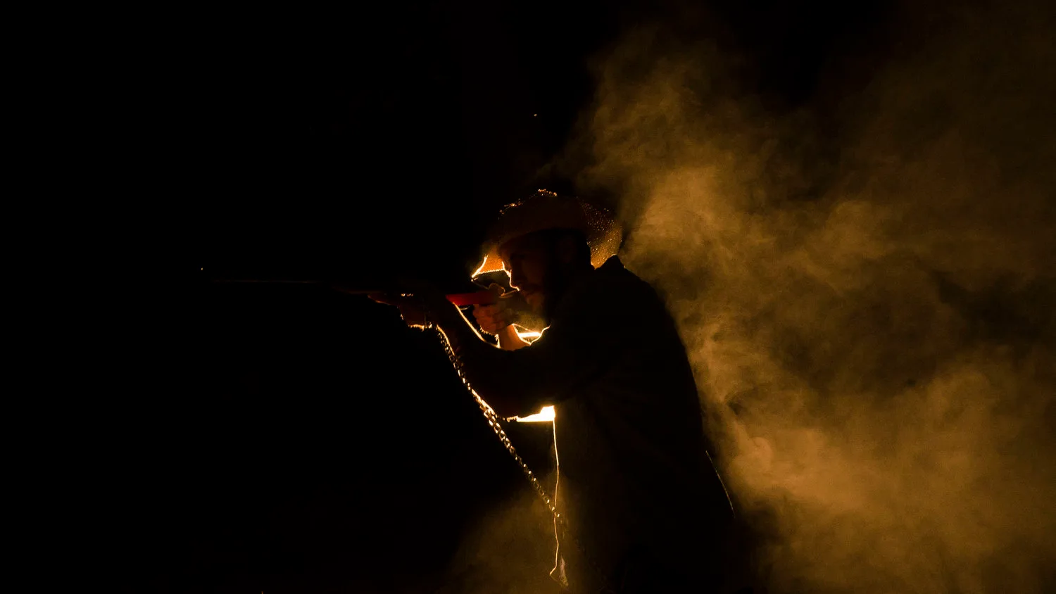 Still from a Rusty Rehl short film: a man in a straw cowboy hat aims a rifle at night, backlit through smoke